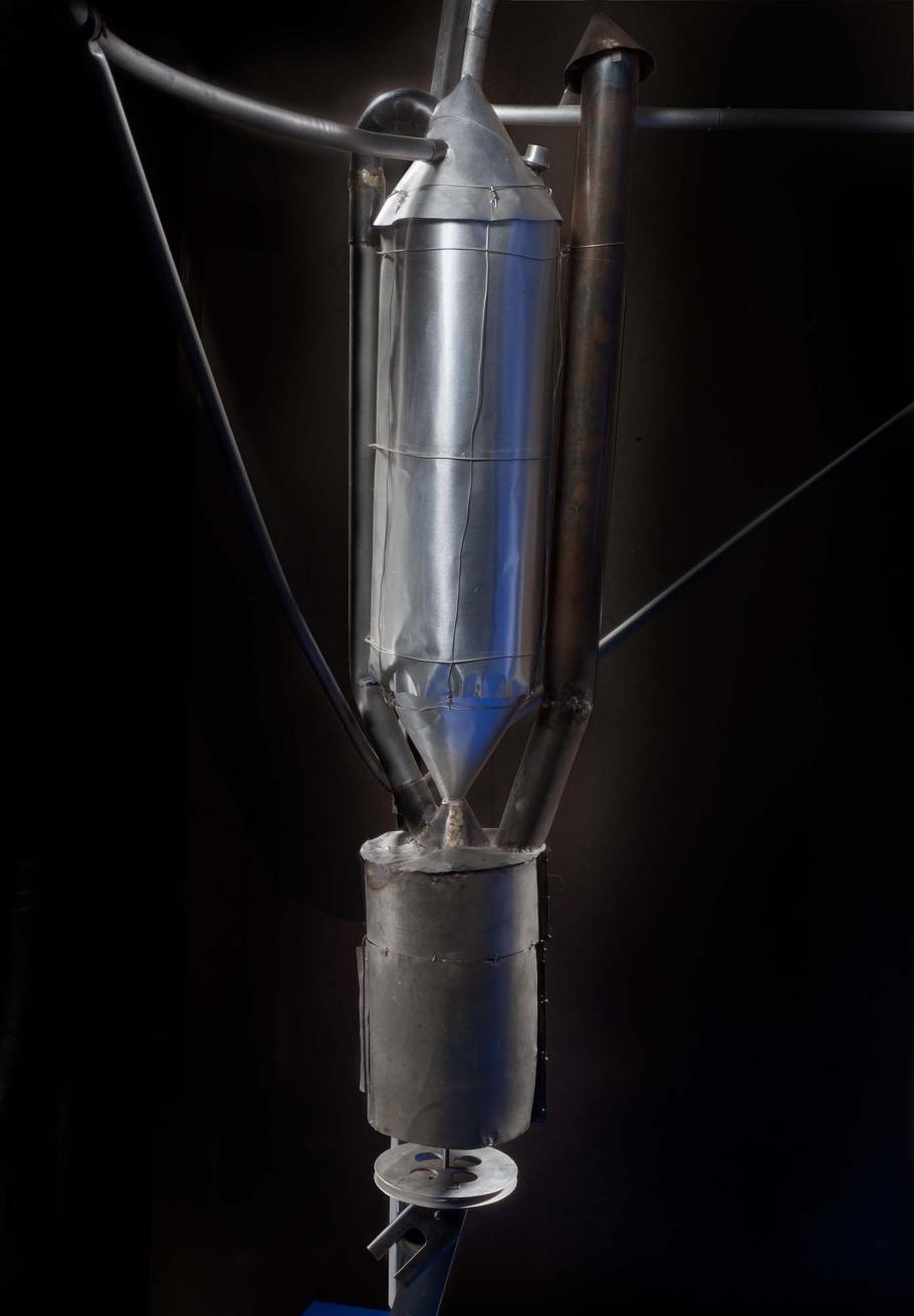 Close-up view of motor on a primitive rocket. A conical nose tops a metal cylinder-shaped motor attached to a cylinder-shaped fuel tank by a metal conical-shaped drain.