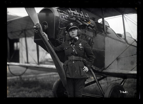 A woman wearing a military or police style uniform, posed beside nose of aircraft. The aircraft is seen in close-up, partial one-half left front view.