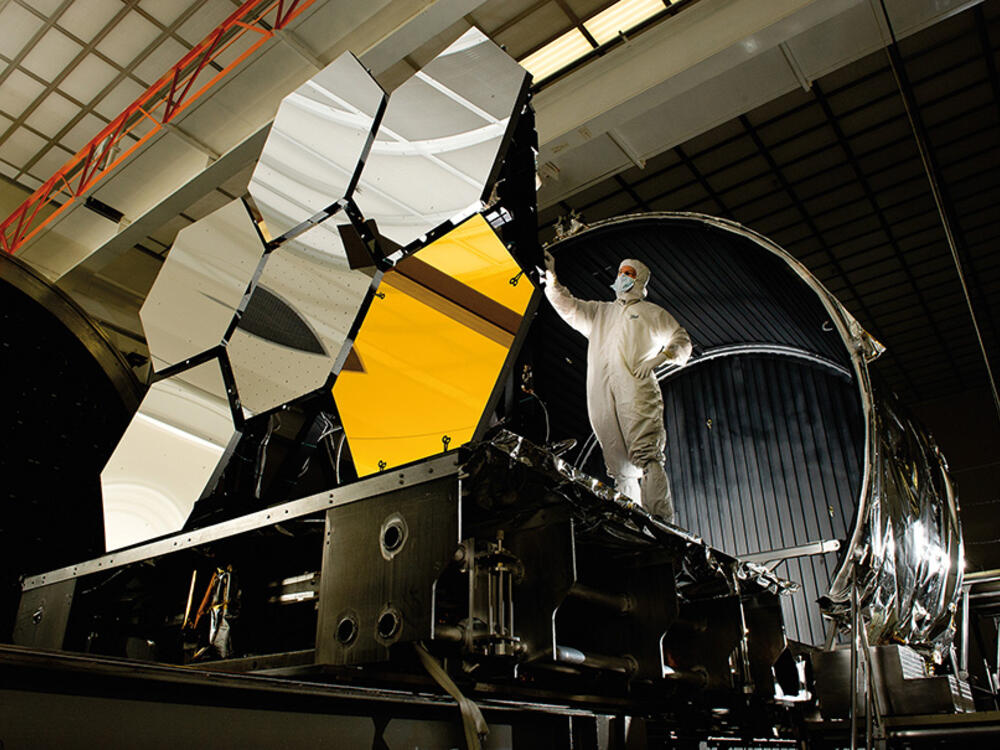 An engineer in a hazmat-type suit and mask inspects the James Webb Space telescope's primary mirror segments, large octagonal mirrors that are the size of the engineer. 