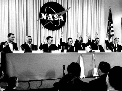 Seven men sit behind a table in front of an audience, each with name cards and microphones in front of them. All seven are raising their hands. 