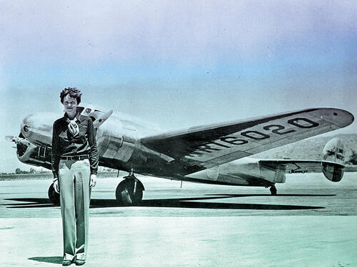A black-and-white portrait of Amelia Earhart standing on an airport tarmac, with her twin-propellered airplane behind her.