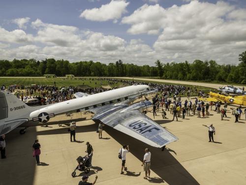 Visitors crowd around historic aircraft outside of the Steven F. Udvar-Hazy Center. In the foreground is a large silver military plane, followed by a smaller yellow one. 